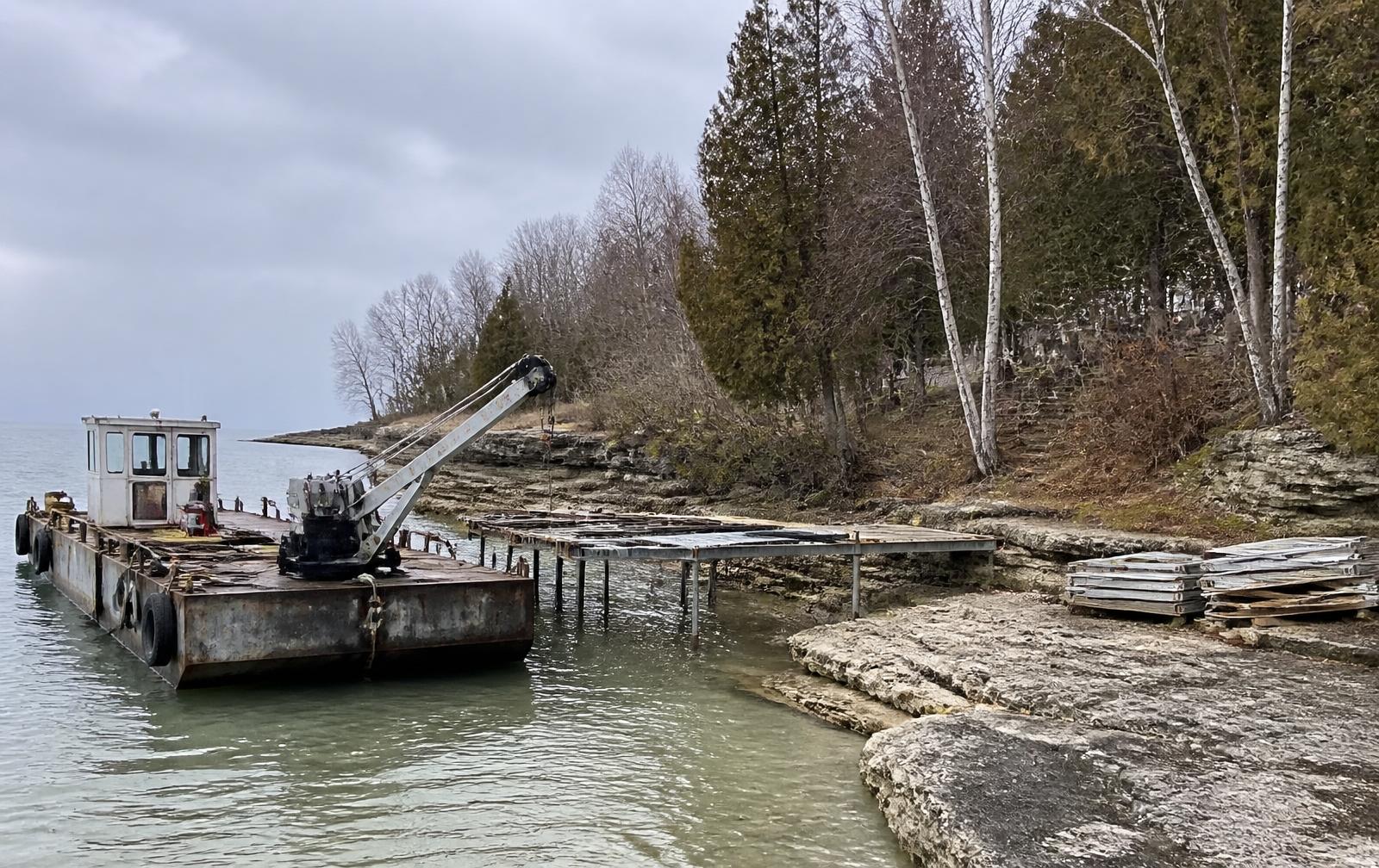 Work barge with a small crane beside a partly removed seasonal pier on a limestone Door County shoreline